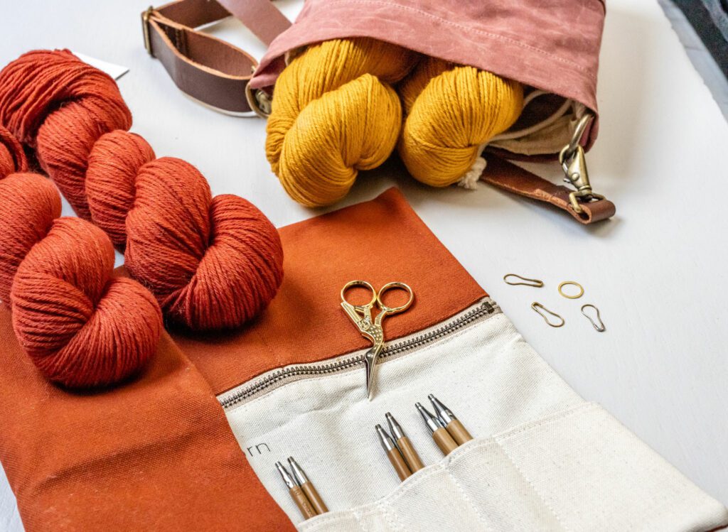 Two red and two curry colored skeins of Rosy Green Wool are on the table.  In addition, you can see an opened needle case from Twig and Horn and the Canvas Bucket Bag.